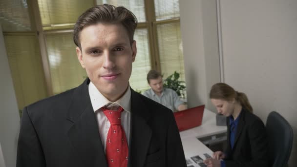 젊은 남자는 양복에서 웃 고는 카메라 보고, 남자와 여자는 배경에서 사무실에서 일하고 있다. 60 fps — 비디오