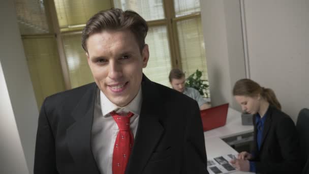 Young man in a suit smiling, shaking his head and looks into the camera, the man and the girl in the background are working in the office. Yes sign, gesture. 60 fps — Stock Video