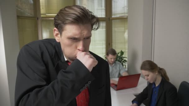 Young man in a suit is coughing, sick, a disease concept. Man and the girl in the background are working in the office. 60 fps — Stock Video