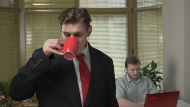 Mladý muž v obleku stojí v úřadu, usmívající se a pít kávu, čaj. Ten pracuje na notebooku v pozadí 60 fps — Stock video