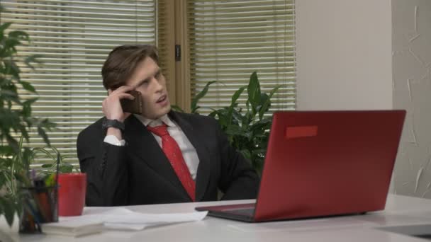 Jeune homme en costume assis au bureau, parlant au téléphone, smartphone, sarcasme, fatigué. Travail dans le concept de bureau 60 fps — Video