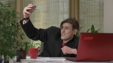 Takım elbiseli genç adam ofiste oturan ve bir selfie yapma. Komik suratlar yapar, bayram, aptallar. Office kavramında işe 60 fps