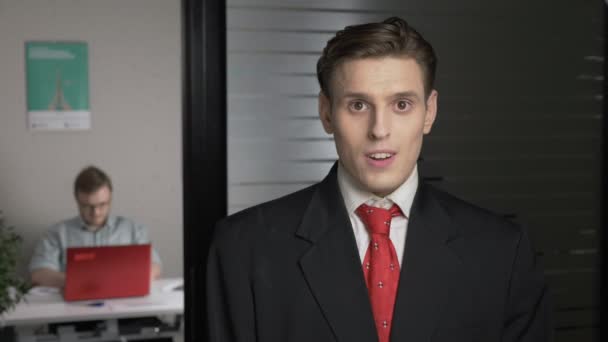 En ung succesfuld mand i jakkesæt viser en følelse af overraskelse, et portræt. Mennesket arbejder på en computer i baggrunden. 60 fps – Stock-video