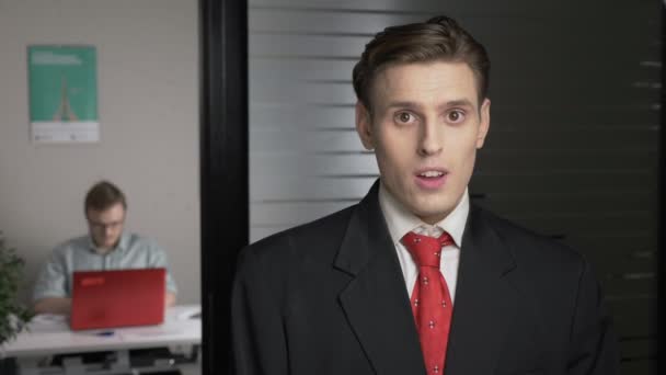 Young successful man in a suit shows an emotion of doubt, uncertain, portrait. Man works on a computer in the background. 60 fps — Stock Video