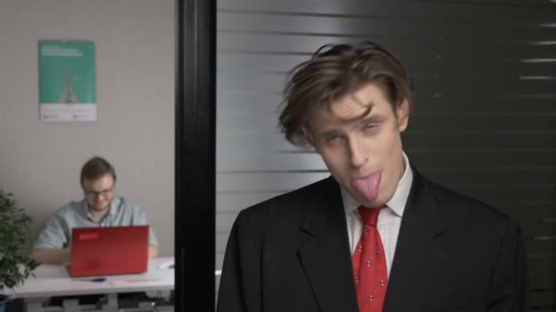 Young successful man in a suit makes funny faces, fools around. Man works on a computer in the background. 60 fps — Stock Video