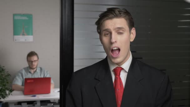 Young successful businessman in a suit bites, clicks his teeth in the office. Man works on a computer in the background. 60 fps — Stock Video