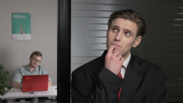 Young successful businessman in a suit thinking, guessing, wondering, have an idea. Man works on a computer in the background. 60 fps — Stock Video