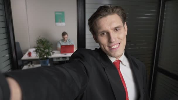 Gros plan de jeune homme d'affaires prospère en costume souriant pour selfies. L'homme travaille sur un ordinateur en arrière-plan. 60 ips — Video