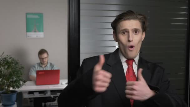 Junger erfolgreicher Geschäftsmann im Anzug zeigt wie Zeichen, Daumen hoch. Mann arbeitet am Computer im Hintergrund. 60 fps — Stockvideo