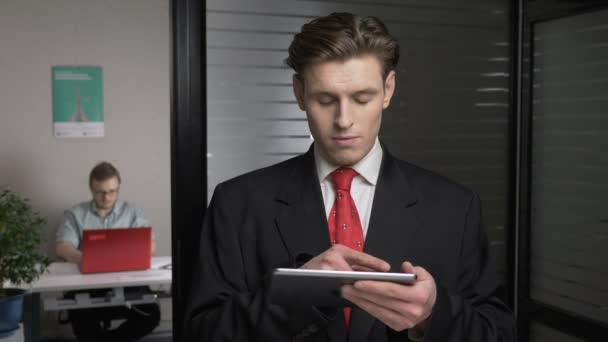 O homem de negócios bem sucedido jovem no terno olha a foto na pastilha, usando uma pastilha. O homem trabalha num computador ao fundo. 60 fps — Vídeo de Stock