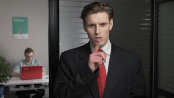 Молодий успішний бізнесмен у костюмі, що показує знак тиші. Людина працює на комп'ютері у фоновому режимі. 60 к/с — стокове відео