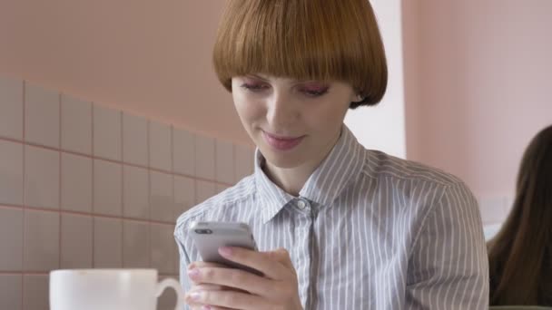 Giovane bella ragazza dai capelli rossi seduta in un caffè, utilizzando uno smartphone, sms, sorridente, ridente. 60 fps — Video Stock