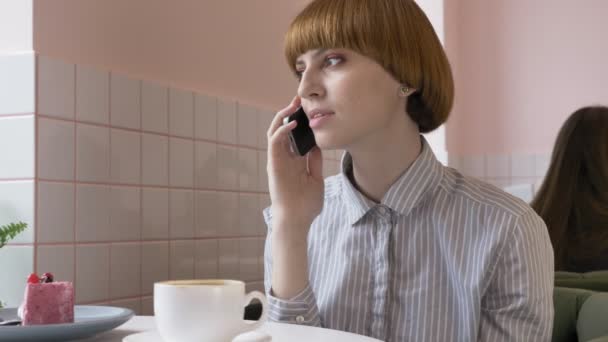 Una joven hermosa pelirroja está sentada en un café, usando un teléfono inteligente, respondiendo a una llamada telefónica, hablando por teléfono. 60 fps — Vídeos de Stock