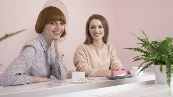 Две молодые кавказские девушки сидят в кафе и улыбаются, глядя в камеру, портрет. Девушки в концепции кафе. 60 кадров в секунду — стоковое видео