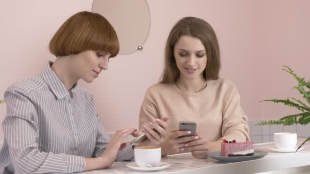 Dos niñas caucásicas jóvenes sentadas en un café y sonriendo, usando un teléfono inteligente, mensajes de texto, mecanografía. Las chicas están en un café. 60 fps — Vídeos de Stock