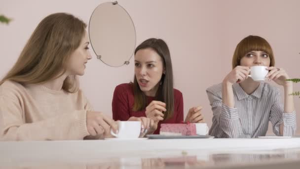 Três jovens caucasianas sentadas em um café, bebendo café, comendo bolo, sorrindo, rindo, gesticulando com as mãos, fofocando velhas namoradas em um conceito de café. 60 fps — Vídeo de Stock