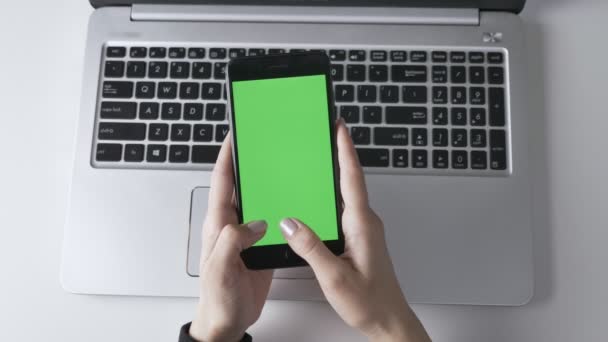 Női kezek gépelés-ra egy nagy fekete smartphone zöld képernyő chroma-key koncepció, laptop, a háttérben. Top shot 60 fps — Stock videók