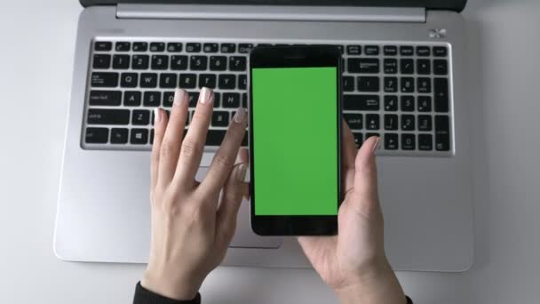 Mani femminili scorrono sullo smartphone touchscreen. Schermo verde, concetto di cromakey, computer portatile in background. Top shot 60 fps — Video Stock
