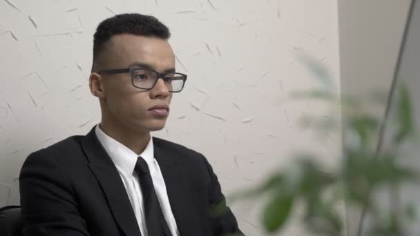 Молодий успішний бізнесмен в окулярах працює на комп'ютері в офісі, посміхається і дивиться на камеру 60 кадрів в секунду — стокове відео