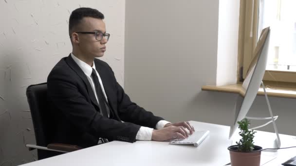 Молодой успешный бизнесмен в очках работает за компьютером в офисе, улыбается и смотрит в камеру 60 кадров в секунду — стоковое видео