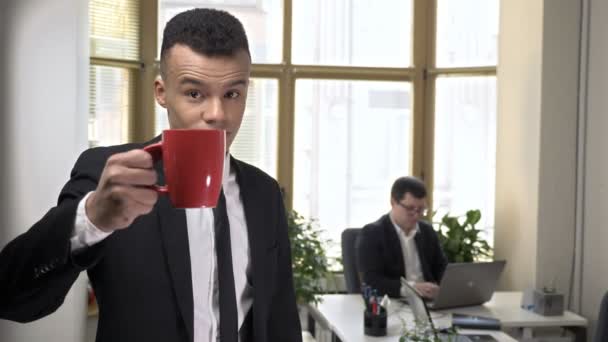 Young successful African businessman drinks coffee from a mug, looking at the camera, portrait, close-up. Man working at the computer in the background. 60 fps — Stock Video