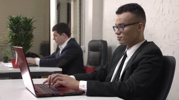 Молодой успешный африканский бизнесмен в очках сидит в офисе и работает на ноутбуке, улыбаясь и глядя в камеру, белый мужчина в костюме на заднем плане. 60 кадров в секунду — стоковое видео
