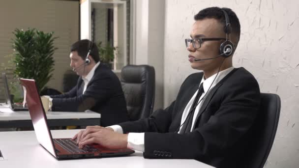 Giovane uomo d'affari africano di successo con gli occhiali si siede in ufficio e lavora sul computer portatile, con auricolare, uomo caucasico in giacca e cravatta sullo sfondo. Concetto di call center. 60 fps — Video Stock