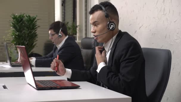 A young successful African man works in the call center, talks on the headset, makes a successful transaction, sells, rejoices in the victory. Caucasian man in suit in the background. Call center — Stock Video