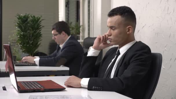 Sekreteri belgeleri alarak cep telefonda konuşurken laptop, ofiste çalışan genç başarılı Afrika erkek iş adamı. Beyaz adam arka planda takım elbiseli. 60 fps — Stok video