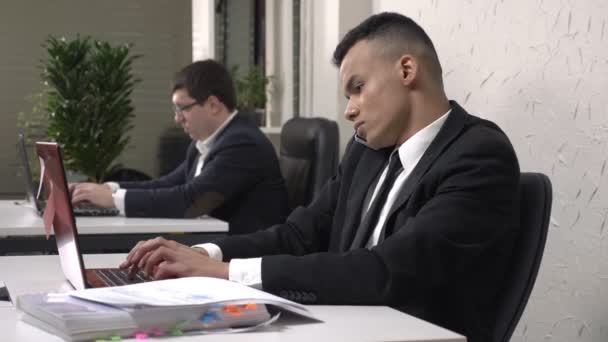 Молодий успішний африканських чоловічого бізнесмен працюючи в офісі на ноутбуку, говорити по мобільному телефону, приймаючи документи від секретаря, дуже зайнятий. 60 кадрів в секунду — стокове відео