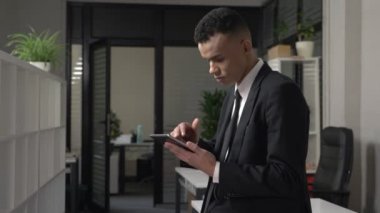 Genç başarılı bir Afrika erkek iş adamı ofis ve tablet, meşgul bir adam bir kostüm kavram kullanma duruyor. 60 fps