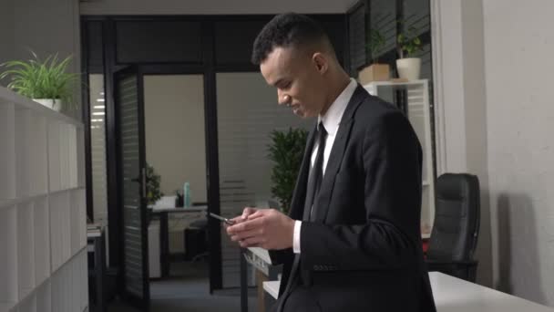 Un joven y exitoso hombre de negocios africano se para en la oficina y usa una tableta, un hombre ocupado en un concepto de vestuario. Sonriendo y jugando en una tableta. Vista lateral. 60 fps — Vídeos de Stock