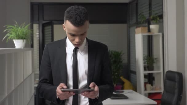 Молодой успешный африканский бизнесмен в костюме стоит в офисе и использует планшет, смс, печатает. Вид спереди. 60 кадров в секунду — стоковое видео