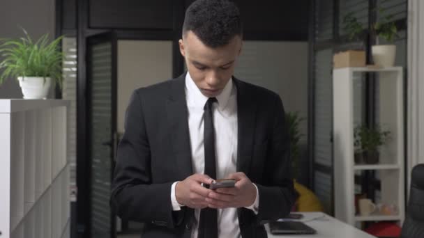 Un joven exitoso hombre de negocios africano con traje se para en la oficina y usa un teléfono inteligente, mensajes de texto, mecanografía. Vista frontal. 60 fps — Vídeos de Stock