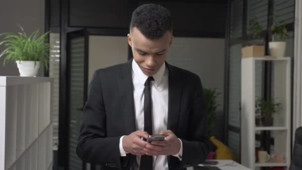 スーツの若い成功したアフリカ男性ビジネスマンがオフィスでスマート フォン、テキスト メッセージ、入力すると、笑みを浮かべて正面を使って立っています。60 fps — ストック動画