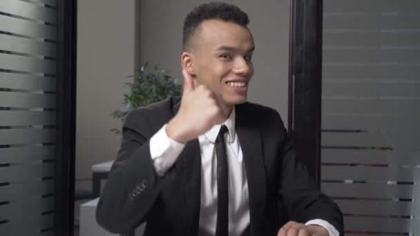 Молодий успішний бізнесмен африканських в костюмі показує знак як сидячи в офісі, посміхаючись. 60 кадрів в секунду — стокове відео