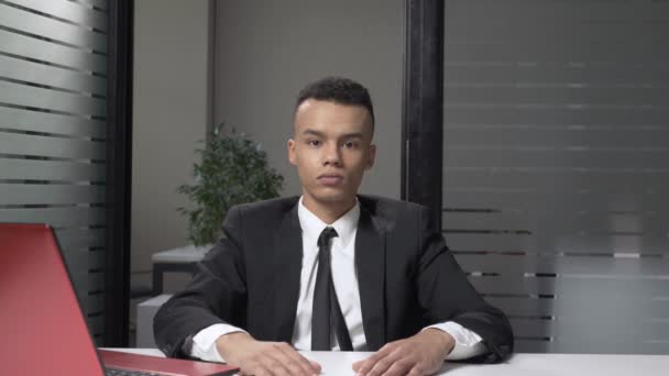 Молодой успешный африканский бизнесмен в костюме показывает знак молчания, сидит в офисе. 60 кадров в секунду — стоковое видео