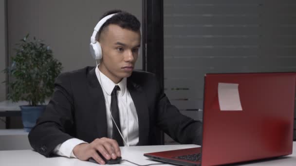 Молодий успішний бізнесмен африканських в костюмі працює на комп'ютері в навушниках, з грою комп'ютер на роботі. 60 кадрів в секунду — стокове відео