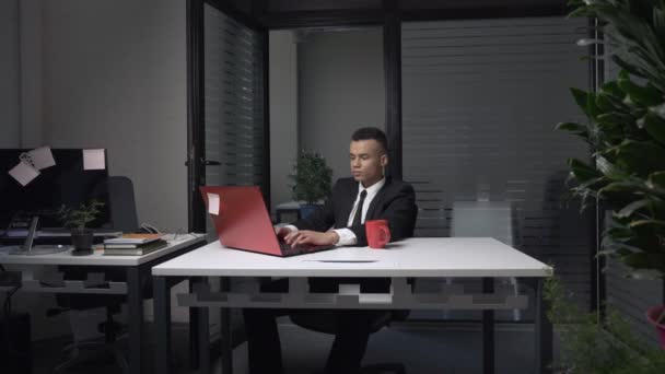 Een jonge succesvolle Afrikaanse zakenman zit in het kantoor, werken op een laptop, koffie drinken uit een beker. 60 fps — Stockvideo