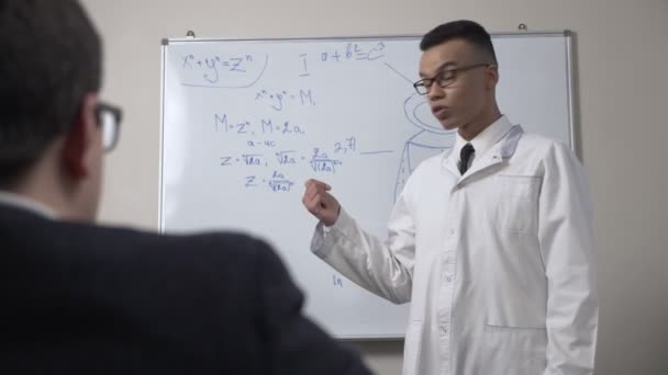 Un giovane scienziato africano di successo in un cappotto bianco e occhiali, si trova alla lavagna con formule, presentazione, lezione, concetto di formazione. 60 fps — Video Stock