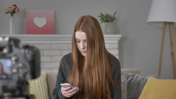 Νεαρή κοκκινομάλλα κοπέλα blogger στο κάμερα, χρησιμοποιώντας ένα smartphone, γραπτών μηνυμάτων, πληκτρολόγηση, σπίτι ζεστασιά στο παρασκήνιο. 60 fps — Αρχείο Βίντεο