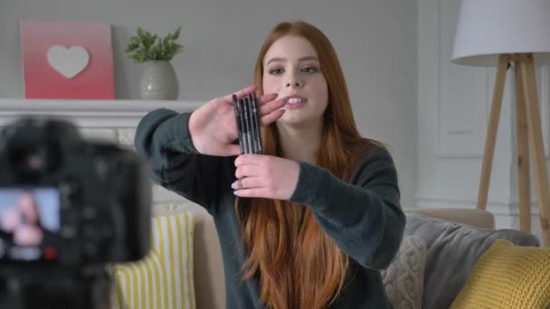 Jonge roodharige meisje blogger, glimlachen, praten op de camera, een nieuwe aankoop, cosmetica, eyeliner, make-up concept, home comfort in de achtergrond tonen. 60 fps — Stockvideo