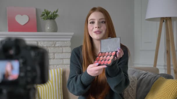 Tânăra blogger cu părul roșu, zâmbind, vorbind la cameră, arătând o nouă achiziție, cosmetice, palet Eyeshadow, confort la domiciliu în fundal. 60 fps — Videoclip de stoc