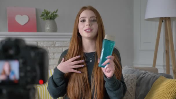 Jeune blogueuse rousse, souriante, parlant à la caméra, montrant un nouvel achat, shampooing, confort à la maison en arrière-plan. 60 ips — Video