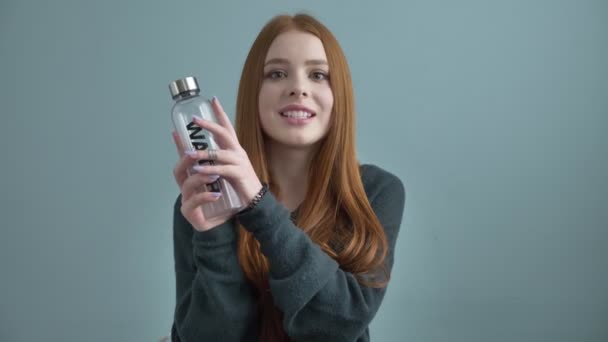 Junge rothaarige Bloggerin, lächelnd, in die Kamera redend, einen Neukauf zeigend, Wasserflasche, Wohnkomfort im Hintergrund. 60 fps — Stockvideo