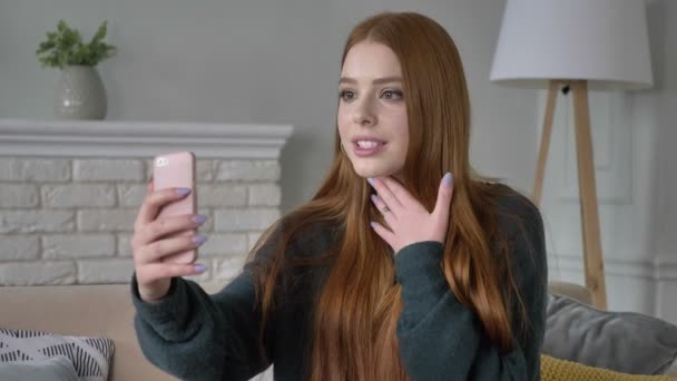 Όμορφη κοκκινομάλλα έφηβος κοπέλα χρησιμοποιεί ένα smartphone, βίντεο chat, άνεση στο σπίτι στο παρασκήνιο. 60 fps — Αρχείο Βίντεο