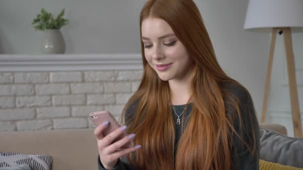 Joven hermosa chica adolescente pelirroja utiliza un teléfono inteligente, sonriendo, mensajes de texto, desplazamiento, deslizamiento, comodidad en el hogar en el fondo. 60 fps — Vídeos de Stock