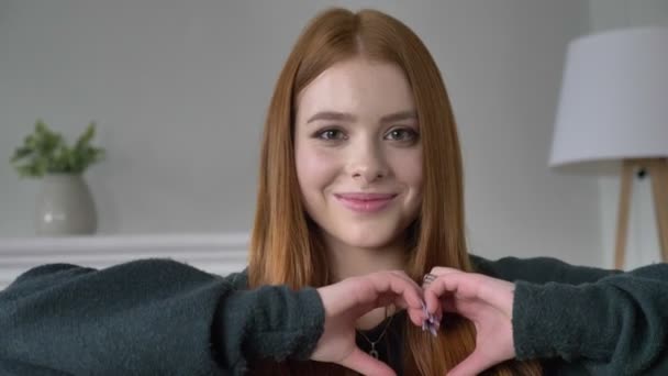 Young red-haired girl blogger, portrait, looking at camera, smiling, showing sign of heart, love, home comfort in the background. 60 fps — Stock Video