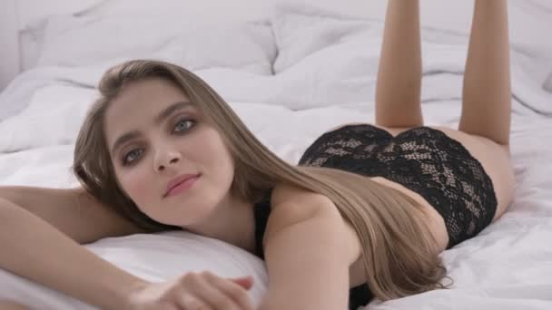 Jonge Kaukasische blond meisje in sexy lingerie ligt op het bed en kijkt naar de camera, portret, verleiding, sexy benen, voeten in de achtergrond, het concept, de 60 fps — Stockvideo