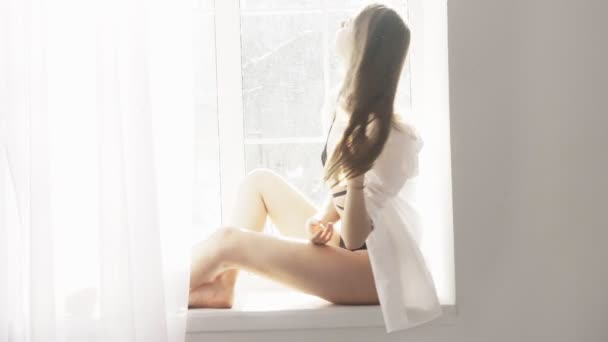 Young beautiful sexy caucasian girl in white shirt and black lingerie sitting by the window, looking at camera, portrait, temptation, sexy, white sunlight in the background. 60 fps — Stock Video